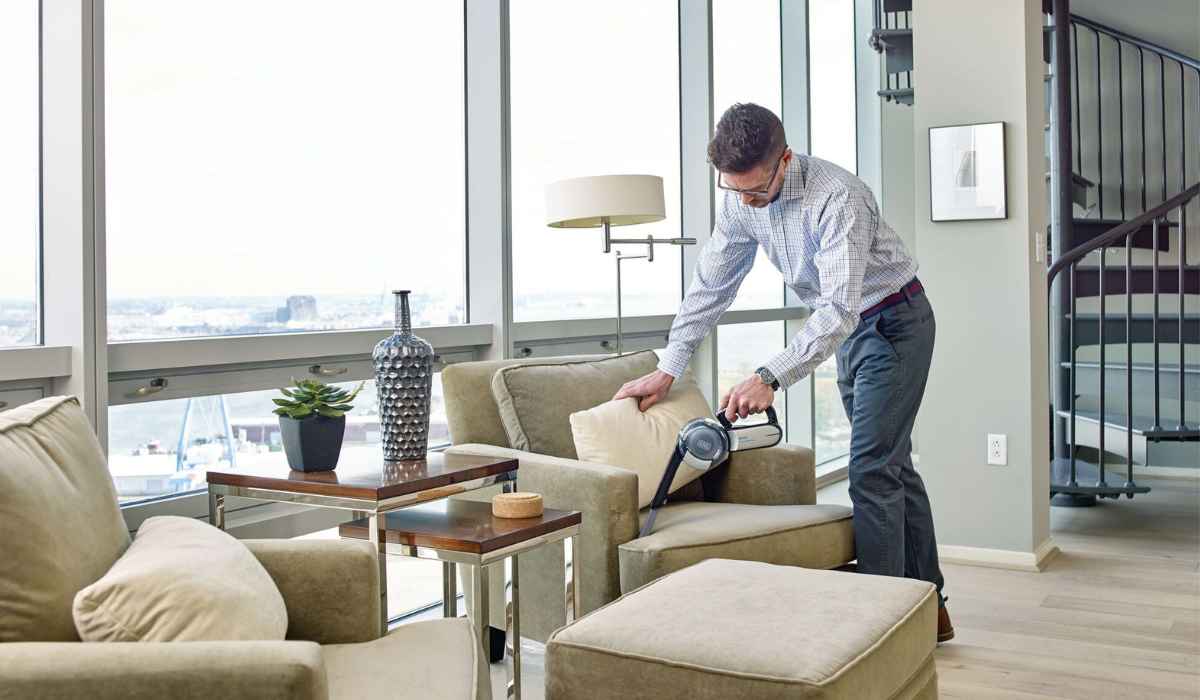 A man using a handheld cordless vacuum to clean a light green armchair in a modern living room with city views.
